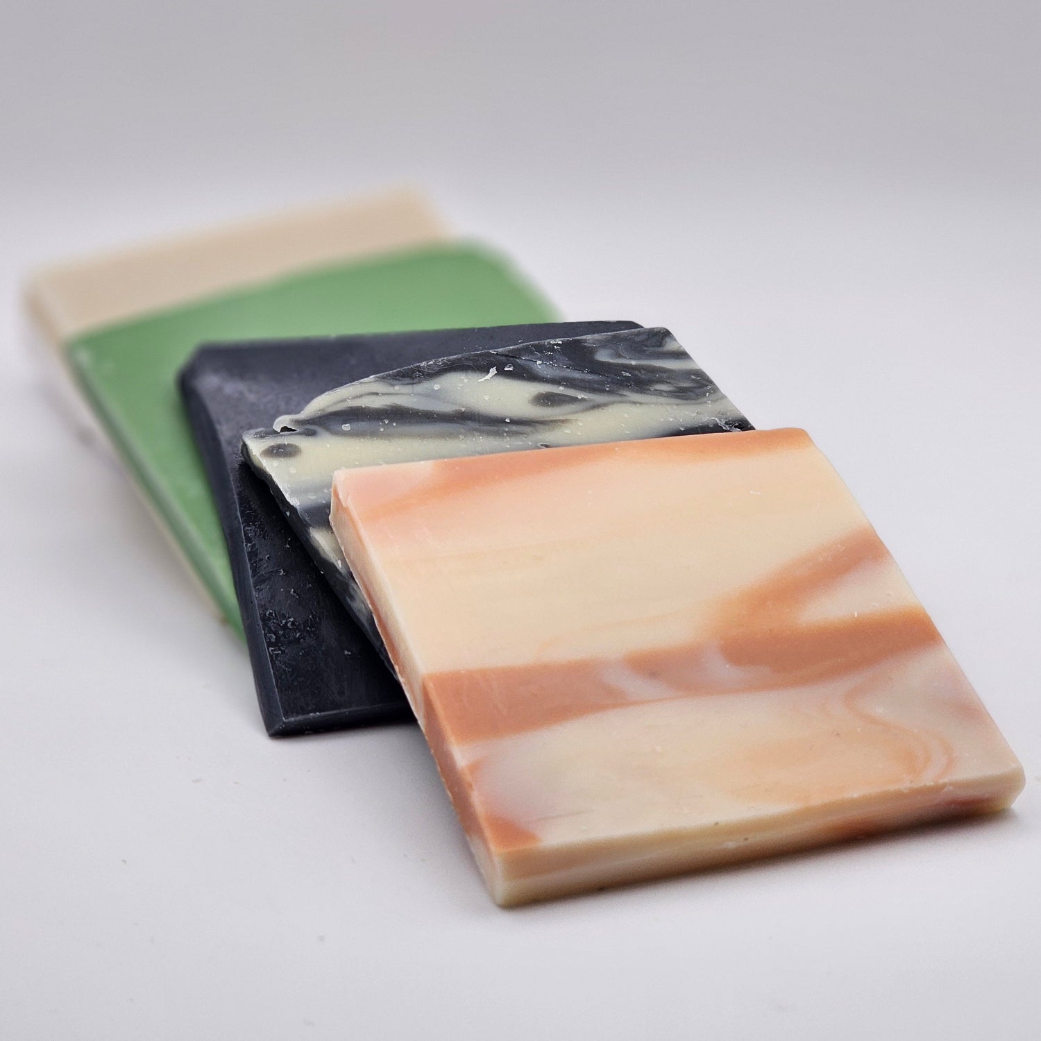 Three rectangular soap bars stacked diagonally on a plain white background. The top soap is marbled cream and peach, the middle is black and white marbled, and the bottom is sage green and cream.